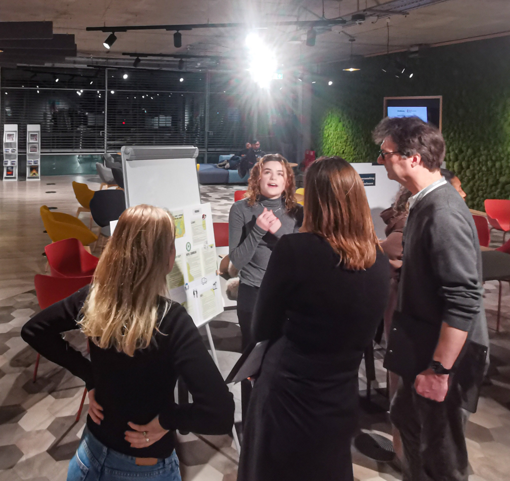 Four people engage in discussion around a flipchart in a modern room. The room features colorful chairs and bright lighting.