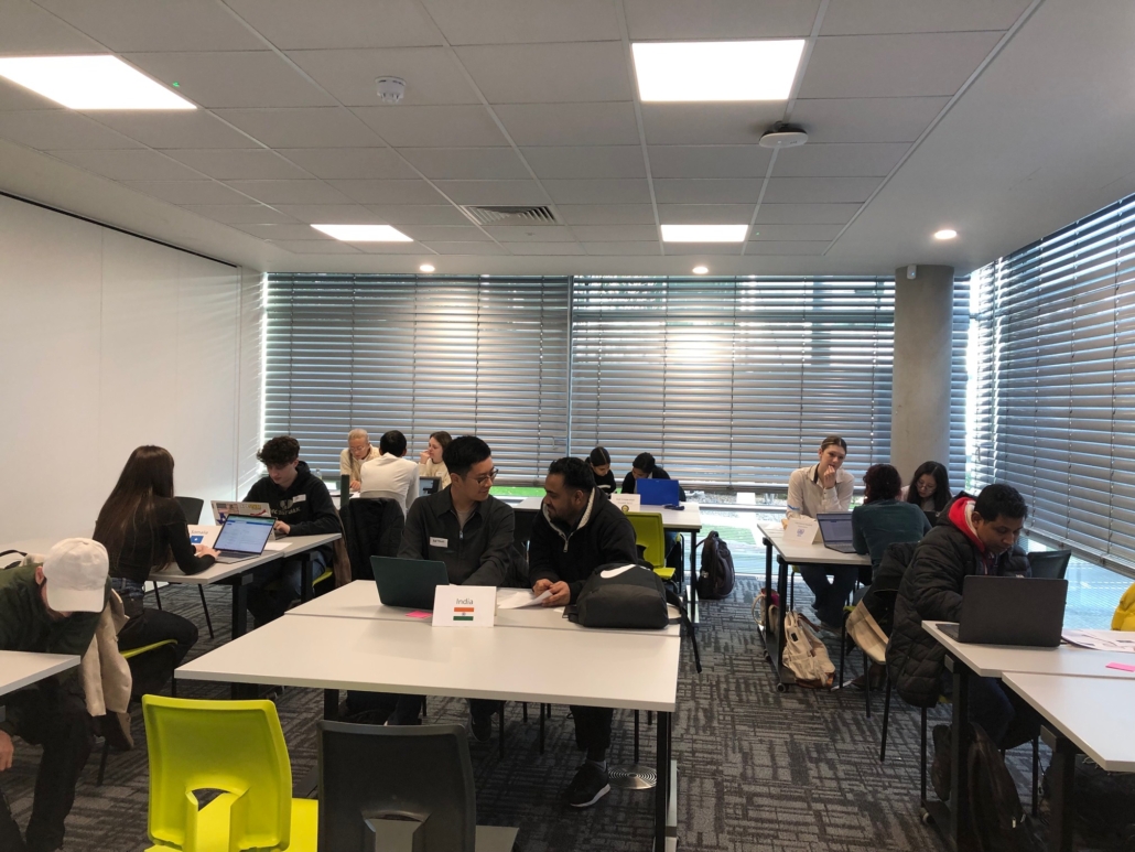 Several people are working on laptops at tables in a modern room with large windows and grid-patterned blinds. Brightly colored chairs are visible.