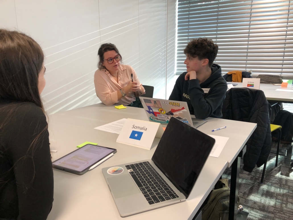 Three people engage in a group discussion around a table with laptops and notes. Signs for Somalia and Eritrea are visible.