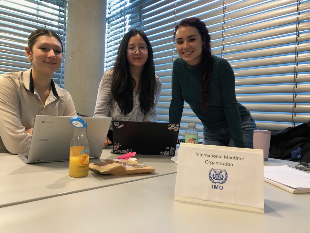 Three people with laptops sit at a table with an International Maritime Organisation sign and bottled drinks, in a room with blinds.