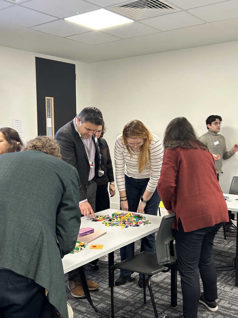 Several people are gathered around a table with colorful Lego pieces in a modern room with white walls and ceiling lights.