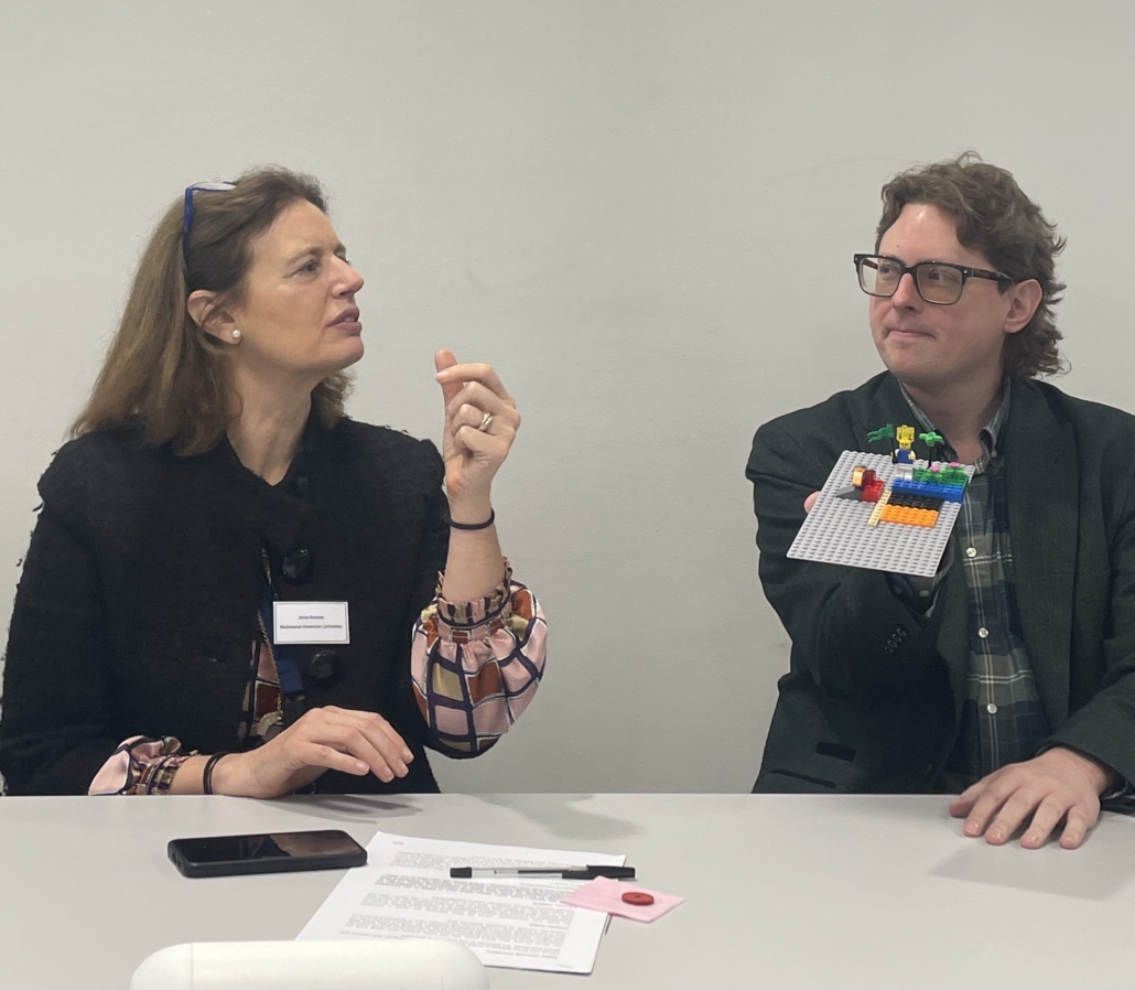 Two people sitting at a table discussing a LEGO model. One person holds the model. Papers and a phone are on the table.