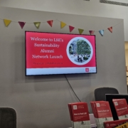 A digital sign announces LSE's Sustainability Alumni Network Launch event. Colorful bunting hangs above with brochures displayed below the screen.