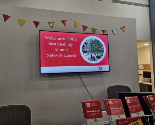 A digital sign announces LSE's Sustainability Alumni Network Launch event. Colorful bunting hangs above with brochures displayed below the screen.
