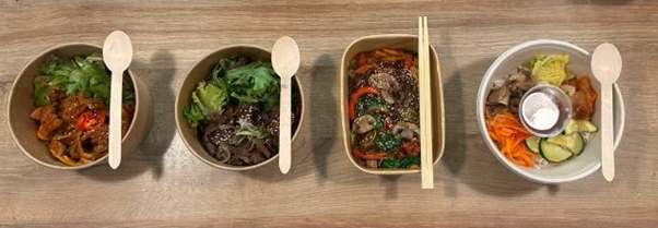 Four bowls of food on a wooden table, each with wooden utensils. Dishes include noodles, vegetables, and meat, presented in an organized manner.