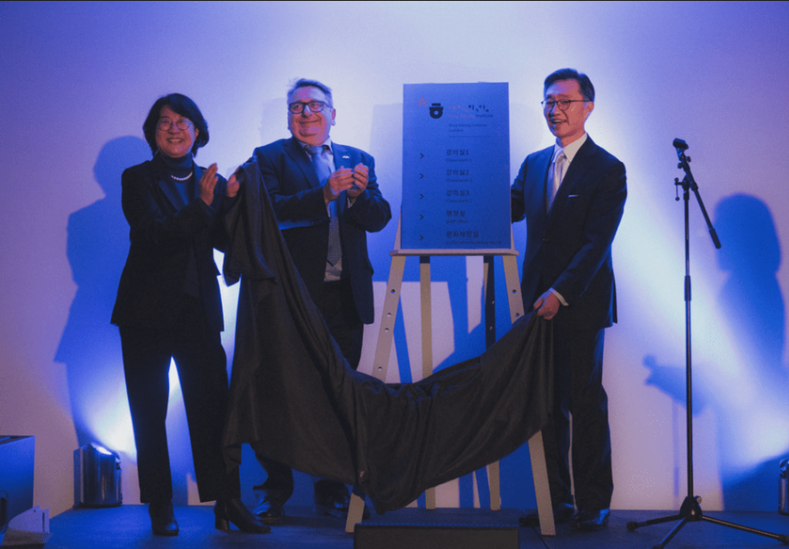 Three people unveiling a plaque on a stand, under blue lighting, with a microphone nearby. Event appears formal and ceremonial.