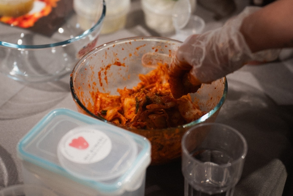 A person prepares kimchi in a bowl, wearing gloves. Various containers and utensils are on the table, suggesting a cooking session.