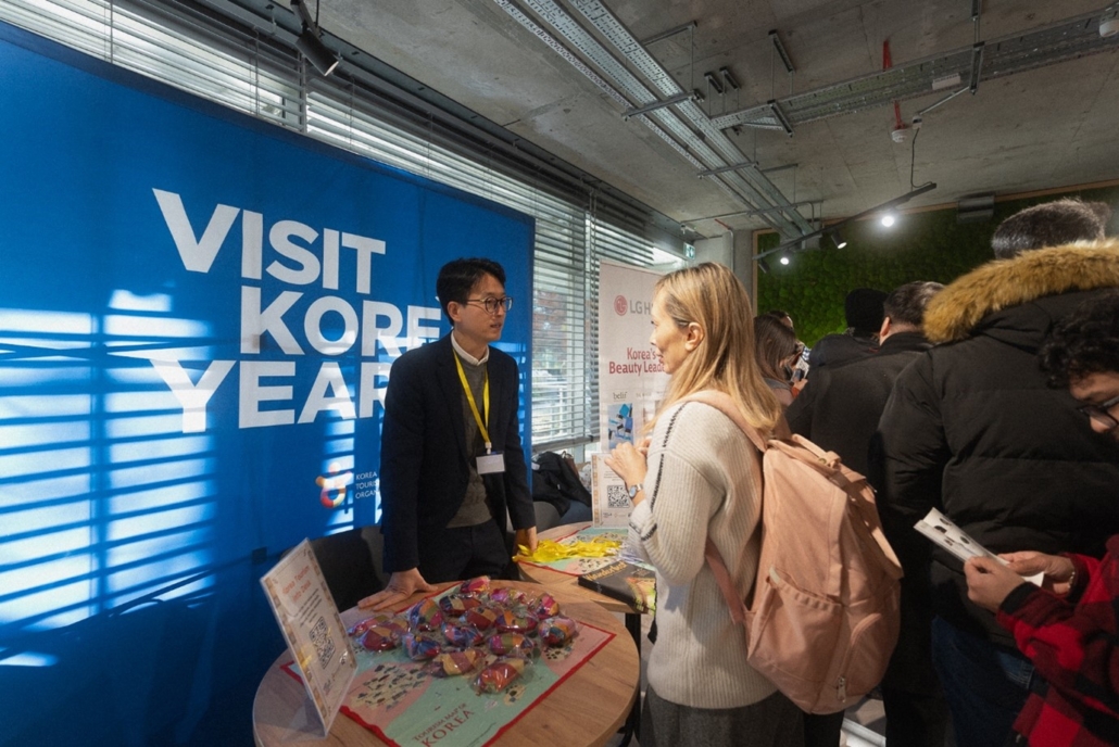 A "Visit Korea Year" promotional booth with people interacting, brochures, and Korean-themed items on display in a modern indoor setting.