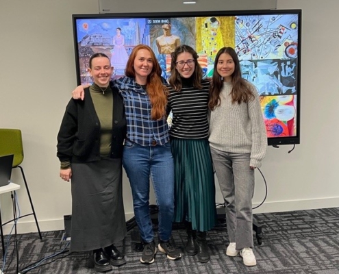 Four people standing together, smiling in front of a large screen displaying various colorful artworks. The room has a modern, professional setting.