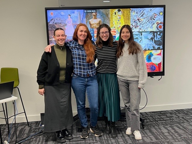 Four people standing together, smiling in front of a large screen displaying various colorful artworks. The room has a modern, professional setting.