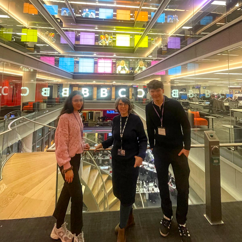 Three people standing inside the BBC Broadcasting House, featuring colorful panels and modern architecture in the background.