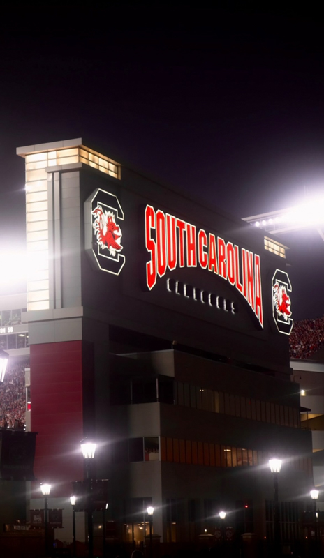Large stadium sign reading "South Carolina" illuminated at night. Gamecock logos visible. Spectators inside the stadium. Stadium lights and tall structure prominent.