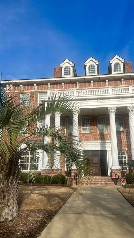 A red-brick building with large white columns and three dormer windows, surrounded by palm trees, under a clear blue sky.
