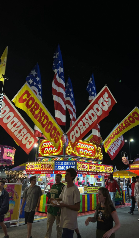 Nighttime carnival scene with people walking. Bright corndog stand adorned with American flags and colorful banners. Festive atmosphere with food and drinks.