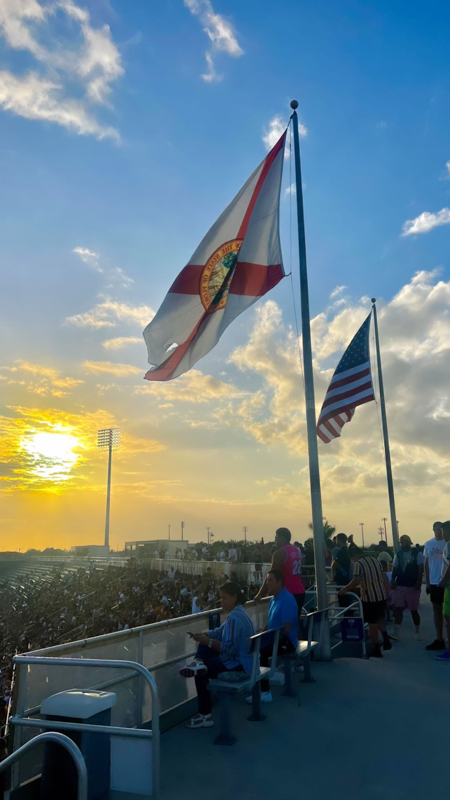 People gather at a stadium during sunset, with the Florida and American flags waving. The sky is partly cloudy and vibrant.