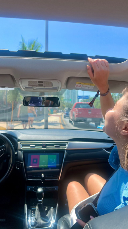 A person adjusts a car's sunroof while parked on a sunny street. Palm trees and buildings are visible through the windshield.