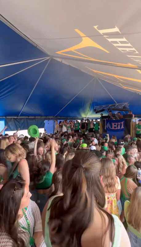 A large, energetic crowd gathers under a blue tent, wearing green attire. A banner with Greek letters is visible in the background.