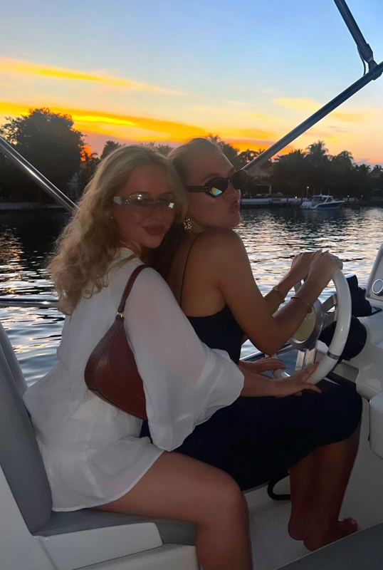 Two people on a boat at sunset, with glasses and casual attire, enjoying a serene waterway with a vibrant sky and surrounding trees.