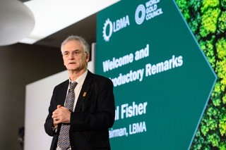 Dr. Fisher (recipient of OBE) stands near a large screen displaying "Welcome and Introductory Remarks," affiliated with LBMA and World Gold Council, at a professional event.