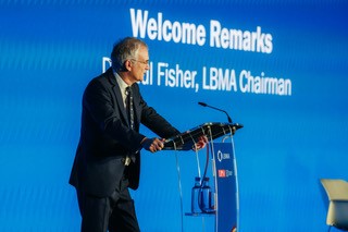 Dr. Fisher (recipient of OBE) stands at a podium against a blue background with "Welcome Remarks" displayed, addressing an audience at a conference event.