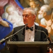 A person in formal attire speaks at a podium. Behind is a large, colorful painting depicting religious figures, including a mother and child.