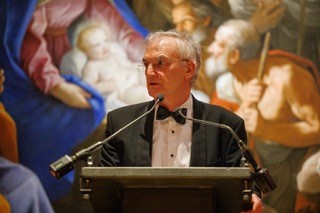 A person in formal attire speaks at a podium. Behind is a large, colorful painting depicting religious figures, including a mother and child.