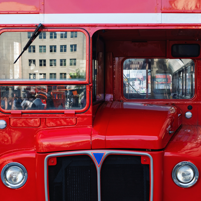 Close-up of a red double-decker bus with reflective windows, showing a city street and several people. An urban building is visible.