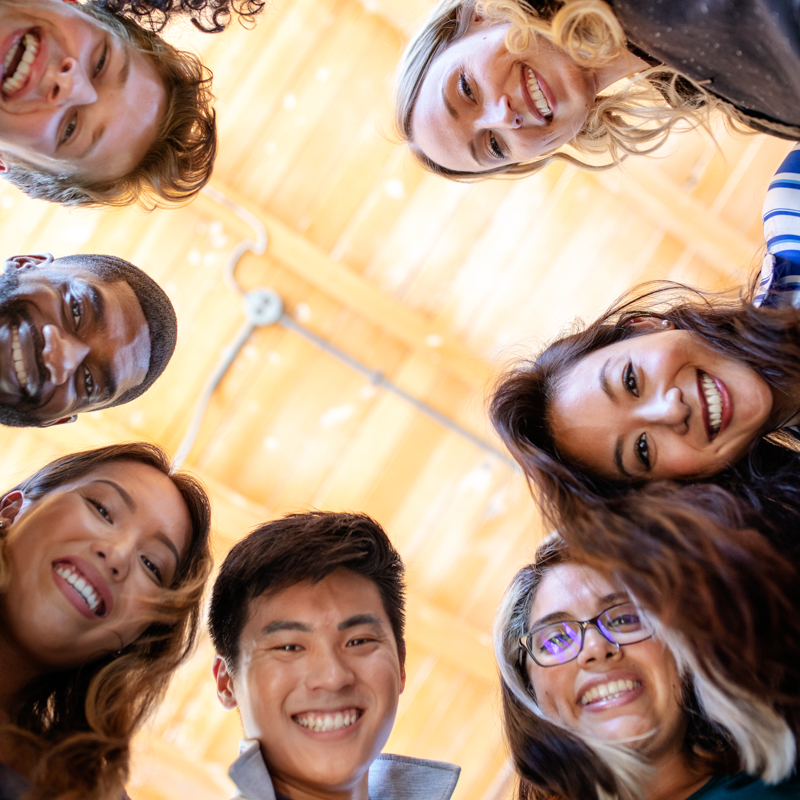 Eight smiling people in a circle, looking down at the camera, set indoors with a wooden ceiling and warm lighting overhead.