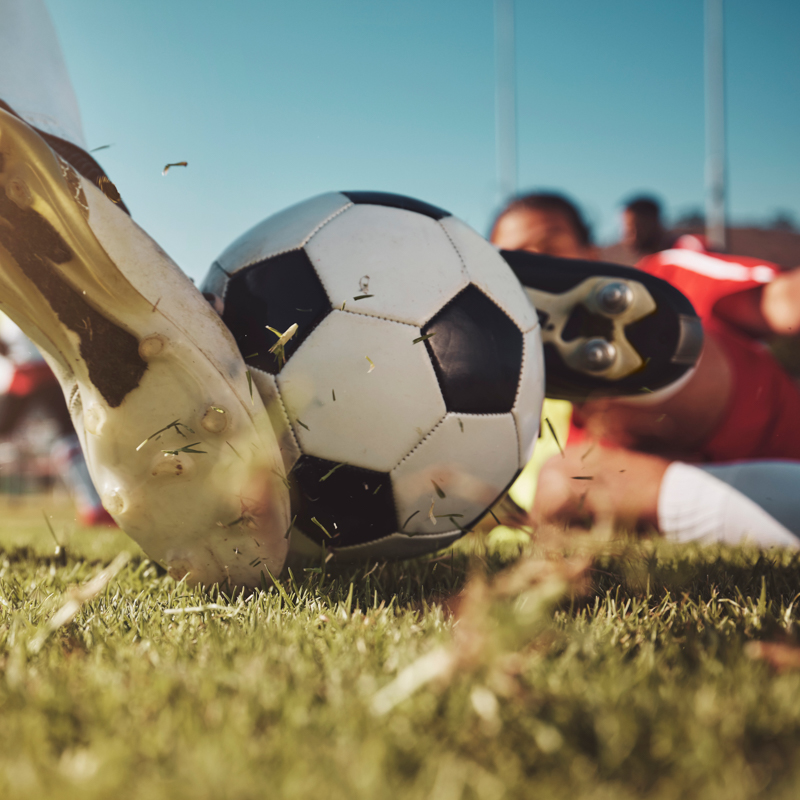 A soccer player in red and another in white cleats compete for the ball on the grassy field, with sky visible above.