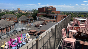 Rooftop terrace with colorful chairs and tables, overlooking a cityscape. Brick buildings and residential area visible under a clear blue sky.