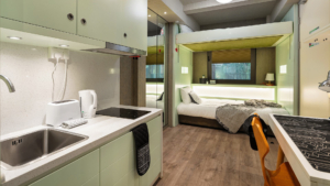 Modern compact studio apartment featuring a small kitchenette, foldable bed, cozy sleeping area, wooden flooring, and contemporary design accents. Sleek and efficient living space.