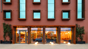 Modern brick building facade with large windows reflecting cityscape, featuring a lit interior and decorative plants on either side of entrance.