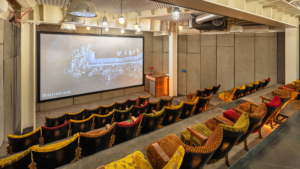 Cozy theater with colorful, vintage-style seating, facing a large screen. A small refreshment area is visible, and industrial lighting hangs overhead.