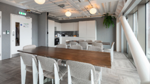 A modern office kitchen with large dining table, chairs, white cabinets, appliances, and plants. Brightly lit interior with wooden flooring.