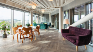 Modern lounge with large windows, wooden floor, and colorful furniture, offering a scenic city view. Natural light enhances the cozy, inviting atmosphere.