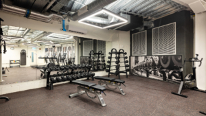 A modern gym features equipment including dumbbells, benches, and exercise bikes in a spacious room with mirrored walls and industrial ceiling design.