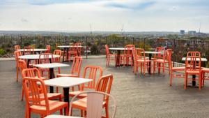 A rooftop terrace with empty orange chairs and tables overlooks a cityscape, featuring distant cranes and buildings under a cloudy sky.