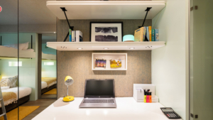 A tidy workspace with a laptop, yellow lamp, books, and personal photos. Bunk beds are reflected in the mirror.
