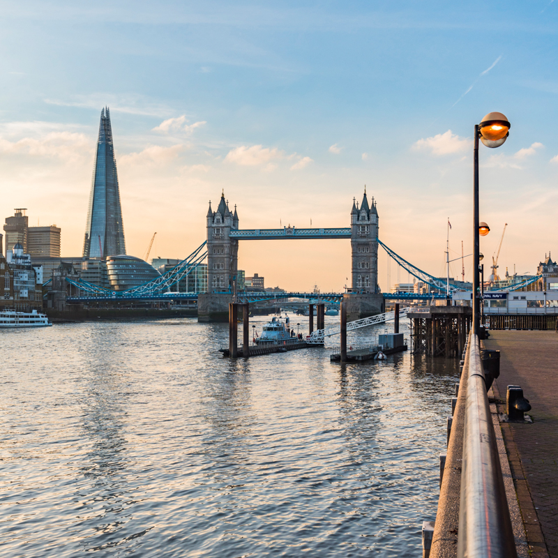 Sunset view of Tower Bridge and The Shard in London, with boats on the Thames River and a riverside walkway.