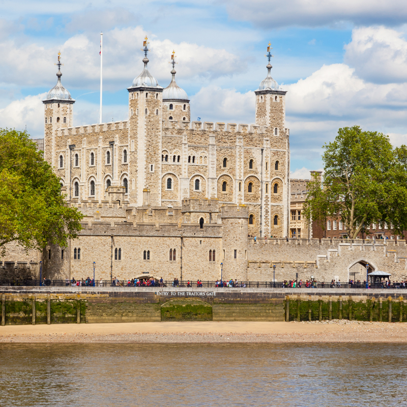 Historic Tower of London by the Thames River, with visitors walking along its walls under a partly cloudy sky and surrounded by greenery.