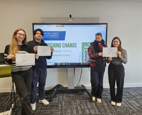 Four people holding certificates stand in front of a presentation screen displaying "John Cabot University" and the words "Engineering Change."