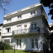 The image shows the United States Embassy in Montevideo, Uruguay, a white multi-story building with a decorative balcony and well-maintained grounds.