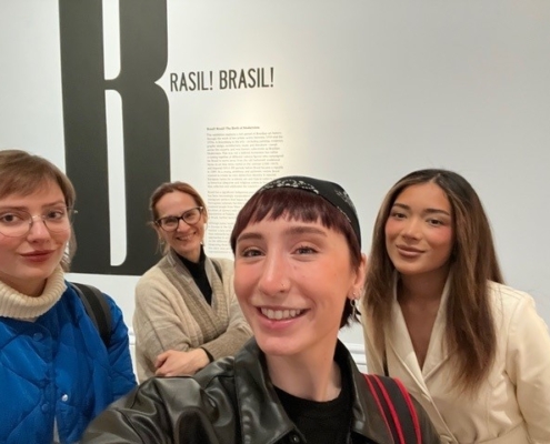 Four people smiling indoors, standing in front of a wall with the text "BRASIL! BRASIL!" No recognizable landmarks or historical buildings visible.