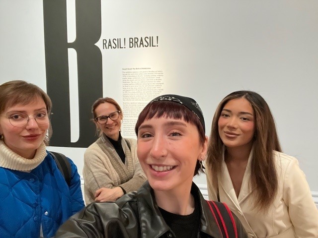 Four people smiling indoors, standing in front of a wall with the text "BRASIL! BRASIL!" No recognizable landmarks or historical buildings visible.