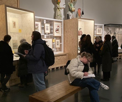 People viewing art in a museum exhibit. One person is sitting on a bench writing. Artwork displayed in wooden and glass cabinets.