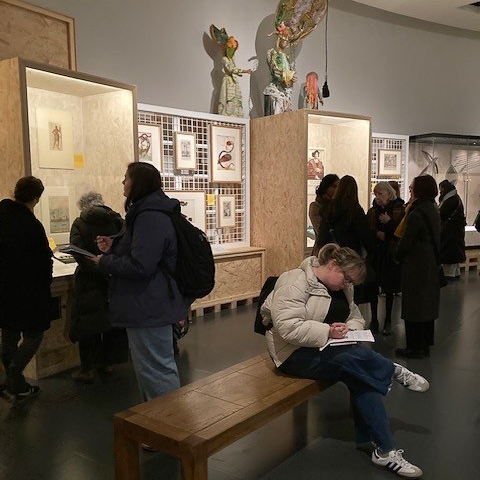 People viewing art in a museum exhibit. One person is sitting on a bench writing. Artwork displayed in wooden and glass cabinets.