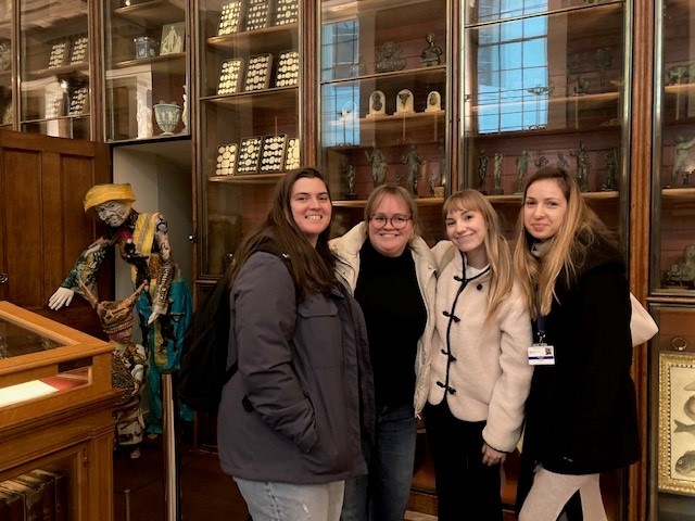 Four people standing in front of cabinets filled with artifacts and figurines, next to a colorful, human-like sculpture in a museum setting.