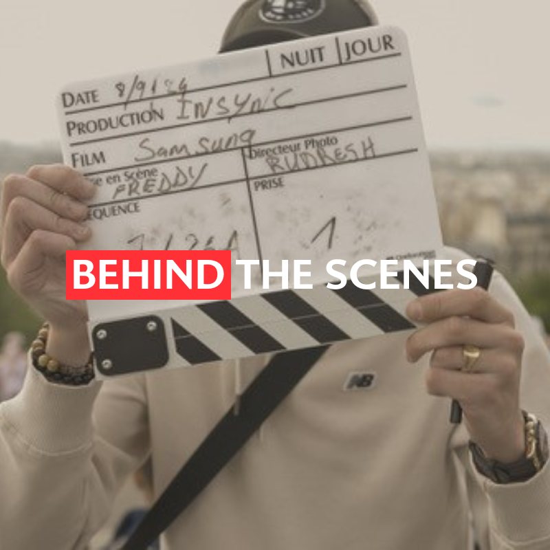 A person holds a clapperboard labeled "Behind the Scenes" in front of a blurred city backdrop.