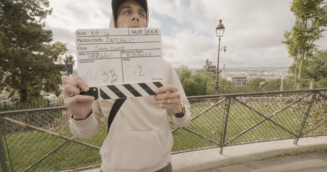 A person holds a film clapperboard outdoors with Paris cityscape in the background, including the Eiffel Tower. Trees and a lamp post are visible.