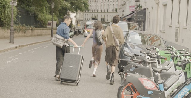 Three people move along a Paris street, one pulling a suitcase. Electric bikes are parked nearby. Trees and buildings line the background.